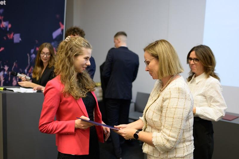 zdjęcie sali, w której odbywa się konferecja. Widać gości i gościnie siedzących przy stołach, a tle siedzą prowadzace spotkanie