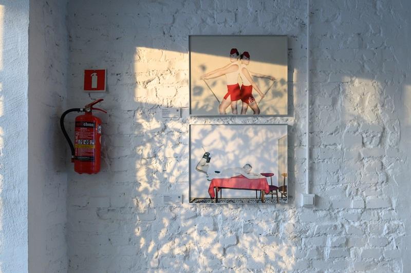 Dwie fotografie autorstwa Rafała Milacha wyeksponowane na białej, ceglanej ścianie. Pierwsze zdjęcie przedstawia dwie mażoretki stojące do siebie plecami, a drugie przedstawia Jana Ślężyńskiego - polskiego profesora nauk o kulturze fizycznej, podczas robienia ćwiczeń na kręgosłup na stole