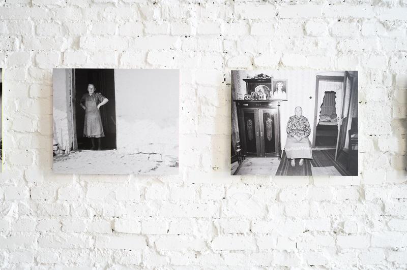 Dwie fotografie autorstwa Zofii Rydet wyeksponowane na ścianie. Na pierwszej widać starszą panią stojącą na ganku, na drugiej kobietę siedzącą na środku pokoju na krześle. W tle widać charakterystyczne koronkowe firanki, a krzesło na którym siedzi stoi na wzorzystej wykładzinie, obok masywnej drewnianej komody. 
