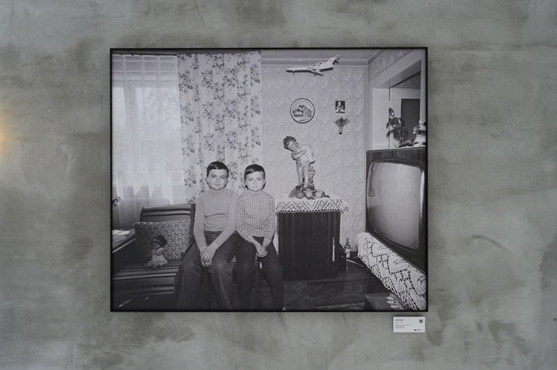Fotografia Zofii Rydet wyeksponowana na ścianie w czarnej ramie. Na zdjęciu widać dwóch chłopców siedzących koło siebie na środku salonu, w którym widać mocno styl lat 70.