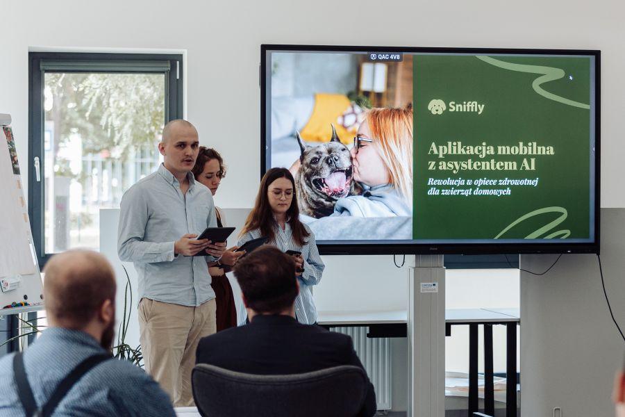 Troje członków zespołu Sniffly prezentuje swój projekt. Na slajdzie zdjęcie psa i napis: „Sniffly. Aplikacja mobilna z asystentem AI. Rewolucja w opiece zdrowotnej dla zwierząt domowych”